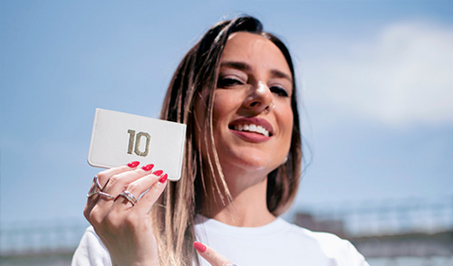 Uma mulher loira sorri para a câmera e segura uma carteira branca com o número 10, licenciada junto ao Santos pela empresa couro50